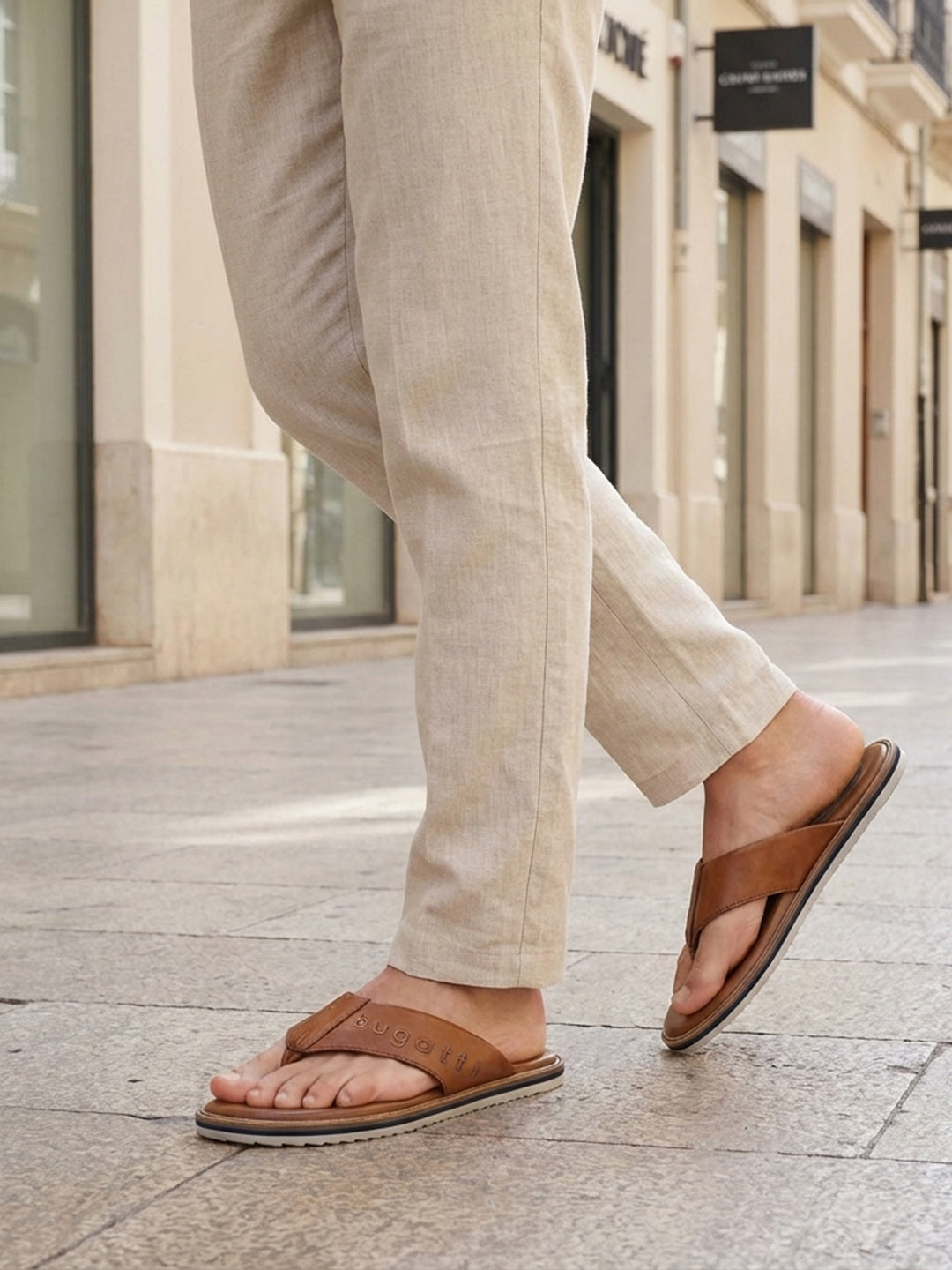 Lifestyle image of bugatti Cobie cognac leather thong sandals worn with casual tan pants in a city setting.