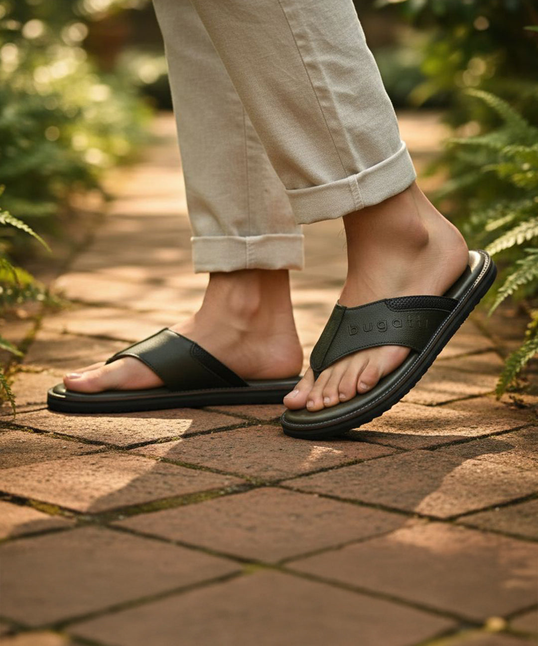 Lifestyle shot of bugatti Cobie dark green leather thong sandals, worn with rolled khaki pants, on a brick path.