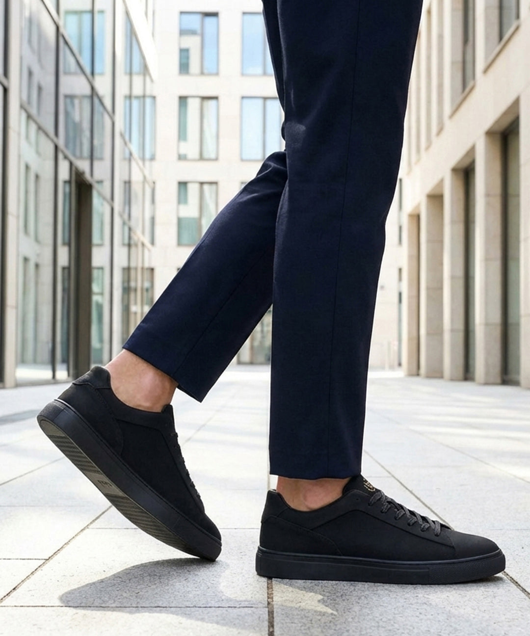 Lifestyle cropped shot of bugatti Fogg Black leather sneakers paired with dark trousers on tiled pavement.