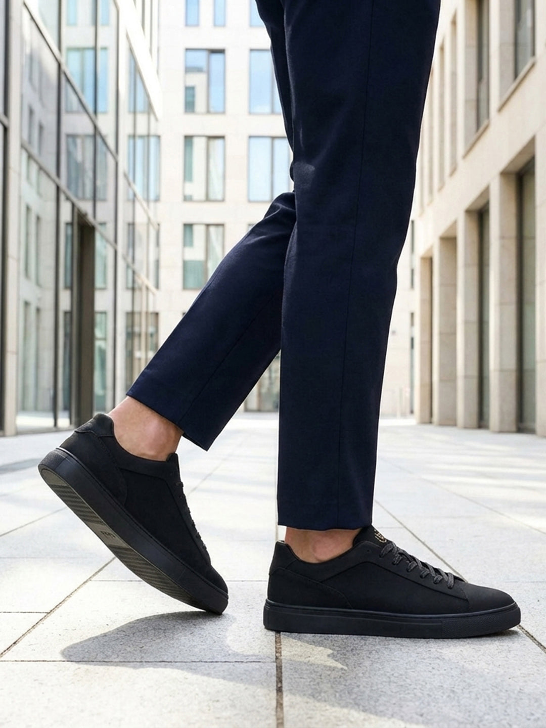 Lifestyle cropped shot of bugatti Fogg Black leather sneakers paired with dark trousers on tiled pavement.