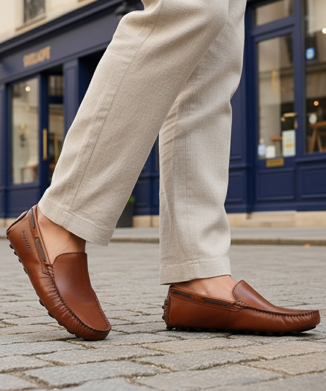 Lifestyle image of bugatti Manfield cognac leather slip-on shoes with visible textured beige pants on a cobblestone street.
