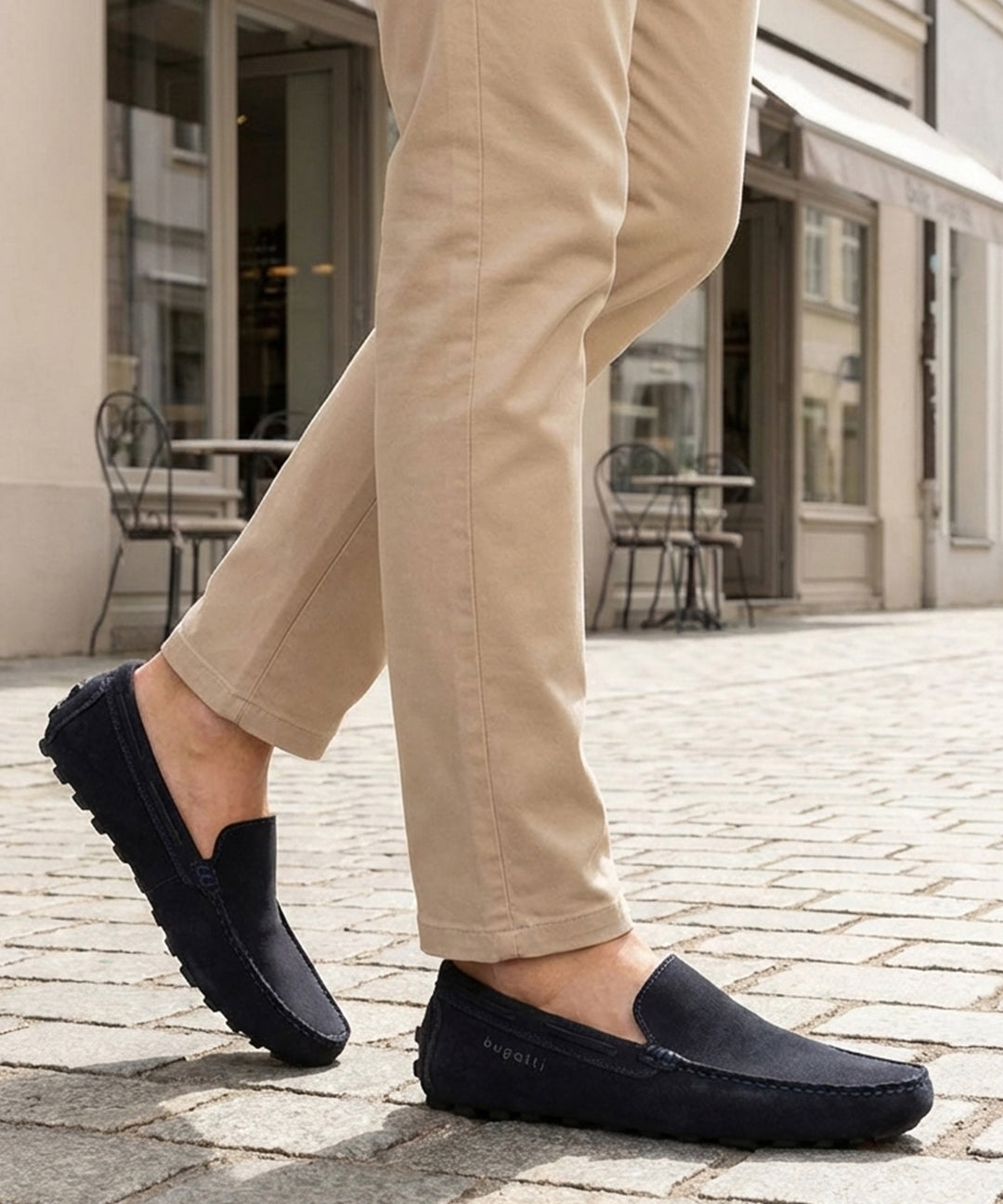 Lifestyle close-up of bugatti Manfield dark blue leather slip-on shoes worn with tan pants on a cobblestone street.
