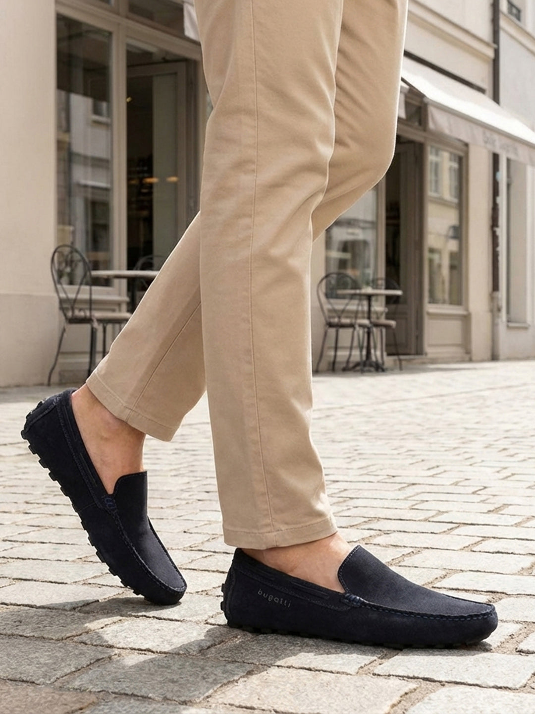 Lifestyle close-up of bugatti Manfield dark blue leather slip-on shoes worn with tan pants on a cobblestone street.
