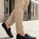 Lifestyle close-up of bugatti Manfield dark blue leather slip-on shoes worn with tan pants on a cobblestone street.
