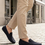 Lifestyle close-up of bugatti Manfield dark blue leather slip-on shoes worn with tan pants on a cobblestone street.

