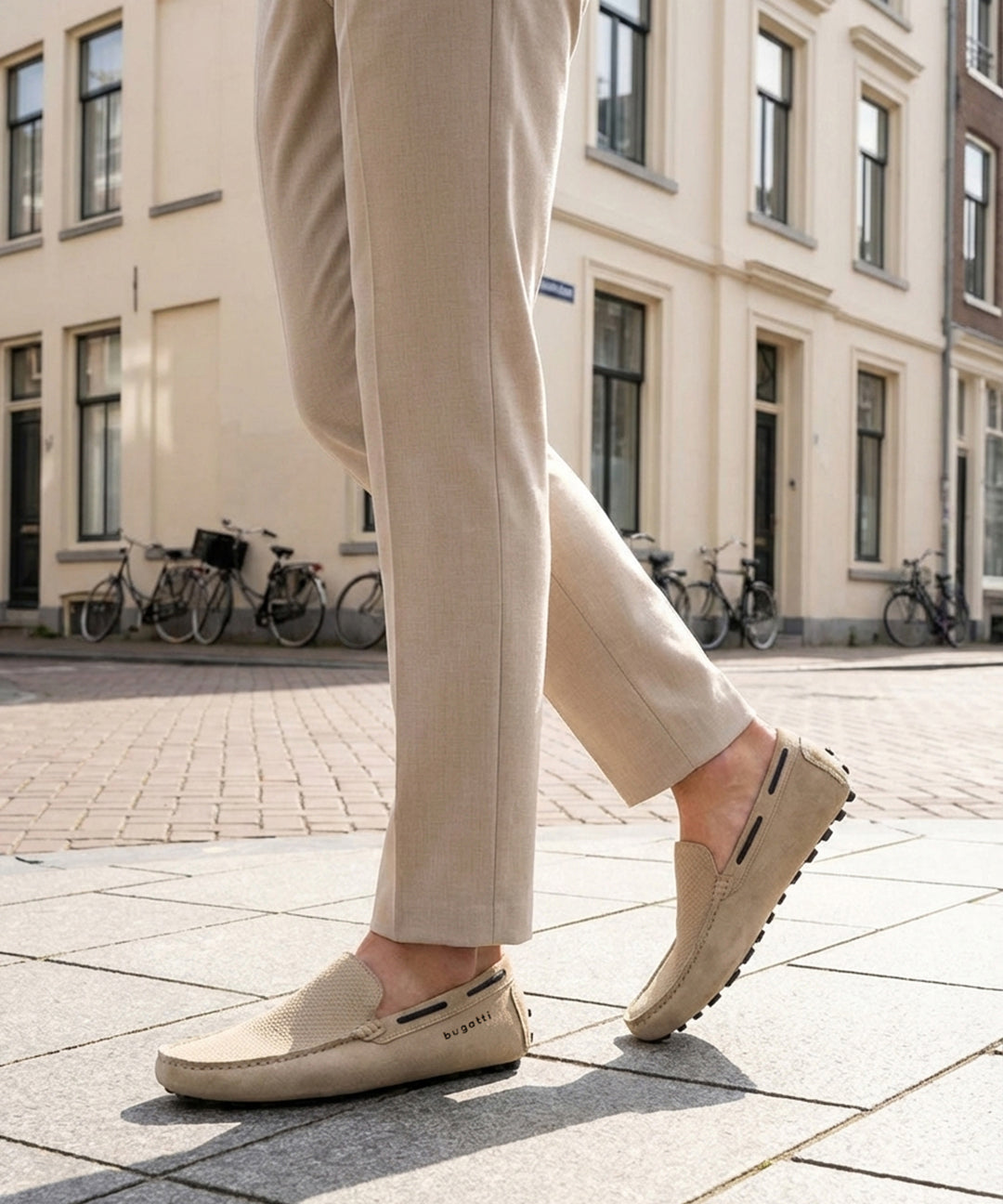 Lifestyle shot of bugatti Manfield sand-colored slip-on shoes in premium leather, worn with tan trousers on a city street.