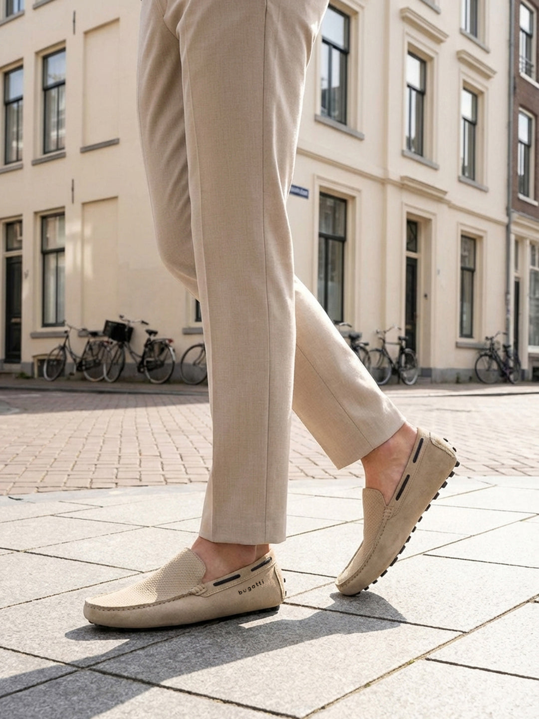 Lifestyle shot of bugatti Manfield sand-colored slip-on shoes in premium leather, worn with tan trousers on a city street.