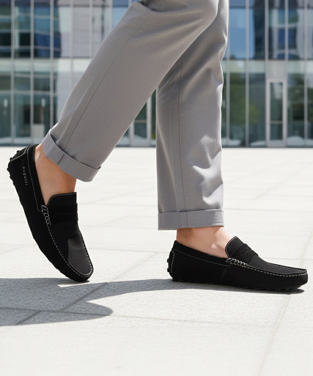 Lifestyle shot of a bugatti Manfield black leather slip-on shoe, paired with gray casual pants.
