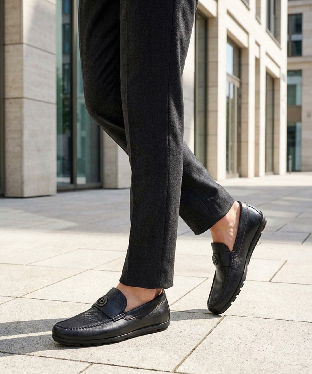 Lifestyle view of black bugatti leather slip-on shoes worn with grey dress pants on a paved surface.
