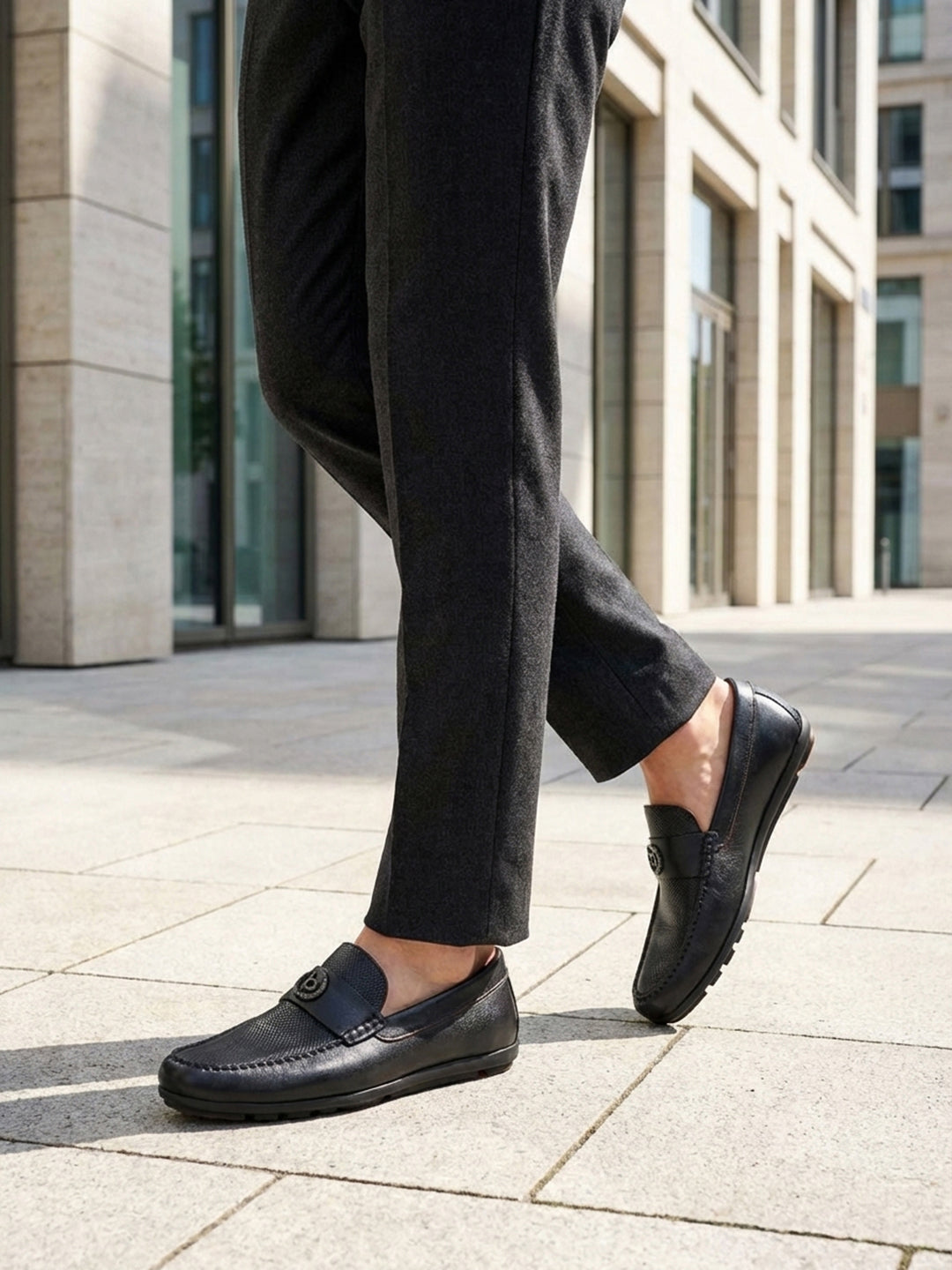 Lifestyle view of black bugatti leather slip-on shoes worn with grey dress pants on a paved surface.