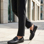 Lifestyle view of black bugatti leather slip-on shoes worn with grey dress pants on a paved surface.