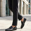 Lifestyle view of black bugatti leather slip-on shoes worn with grey dress pants on a paved surface.