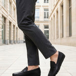 bugatti Cobe black premium leather slip-on shoe, side view, paired with gray dress pants in a city setting.
