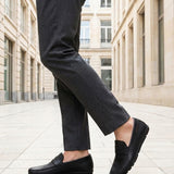 bugatti Cobe black premium leather slip-on shoe, side view, paired with gray dress pants in a city setting.
