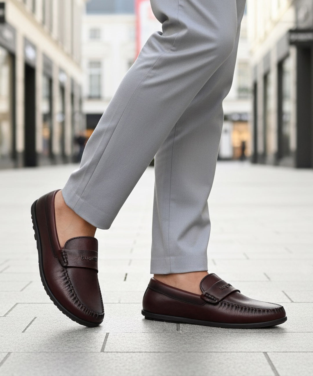 Lifestyle shot of a bugatti Linex slip-on shoe in mid-brown leather, paired with gray slacks and worn on a tiled street.

