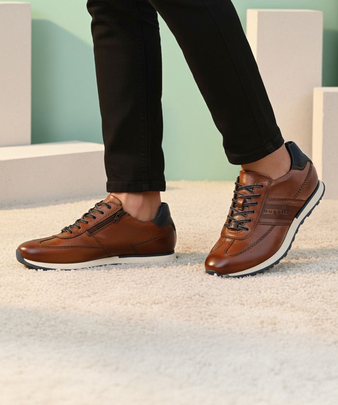 Lifestyle shot of brown leather bugatti sneakers with black laces and dark blue accent, worn with black pants.
