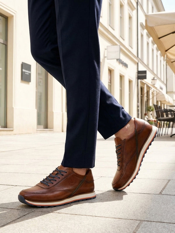 Lifestyle image of bugatti Garnet Evo cognac leather sneakers with white soles, worn with dark blue pants.