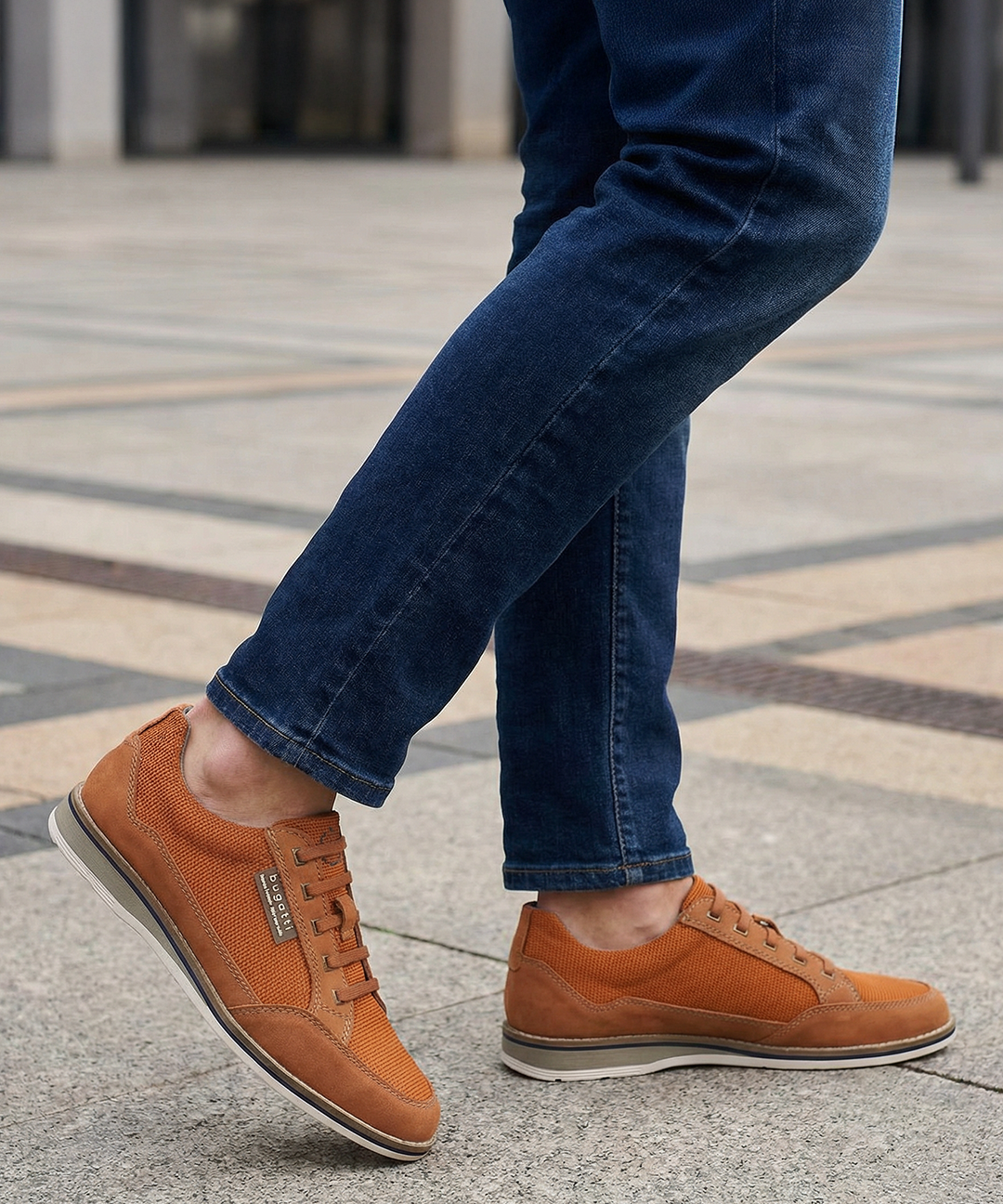 Cropped shot of a person wearing bugatti Makkor orange casual shoes with blue jeans on a concrete surface.
