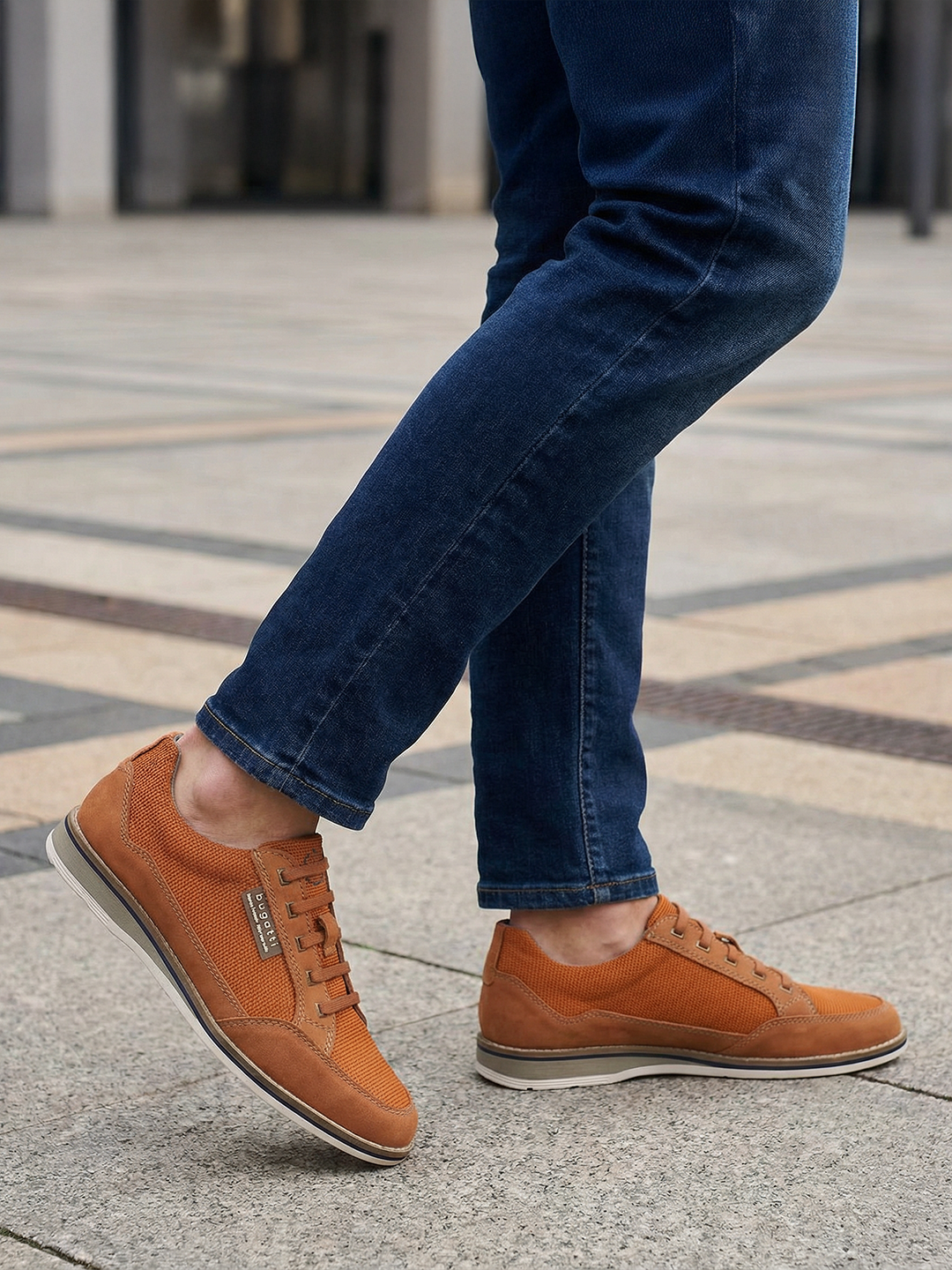 Cropped shot of a person wearing bugatti Makkor orange casual shoes with blue jeans on a concrete surface.
