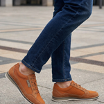 Cropped shot of a person wearing bugatti Makkor orange casual shoes with blue jeans on a concrete surface.

