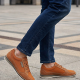 Cropped shot of a person wearing bugatti Makkor orange casual shoes with blue jeans on a concrete surface.
