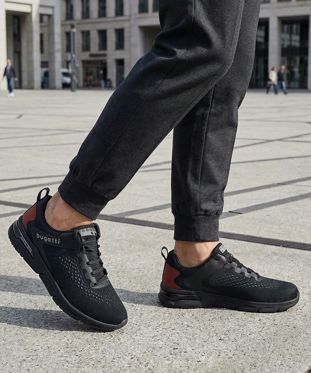 Lifestyle close-up of black bugatti Nubola comfort sneakers, with mesh texture and red heel accent, worn with dark gray sweatpants.
