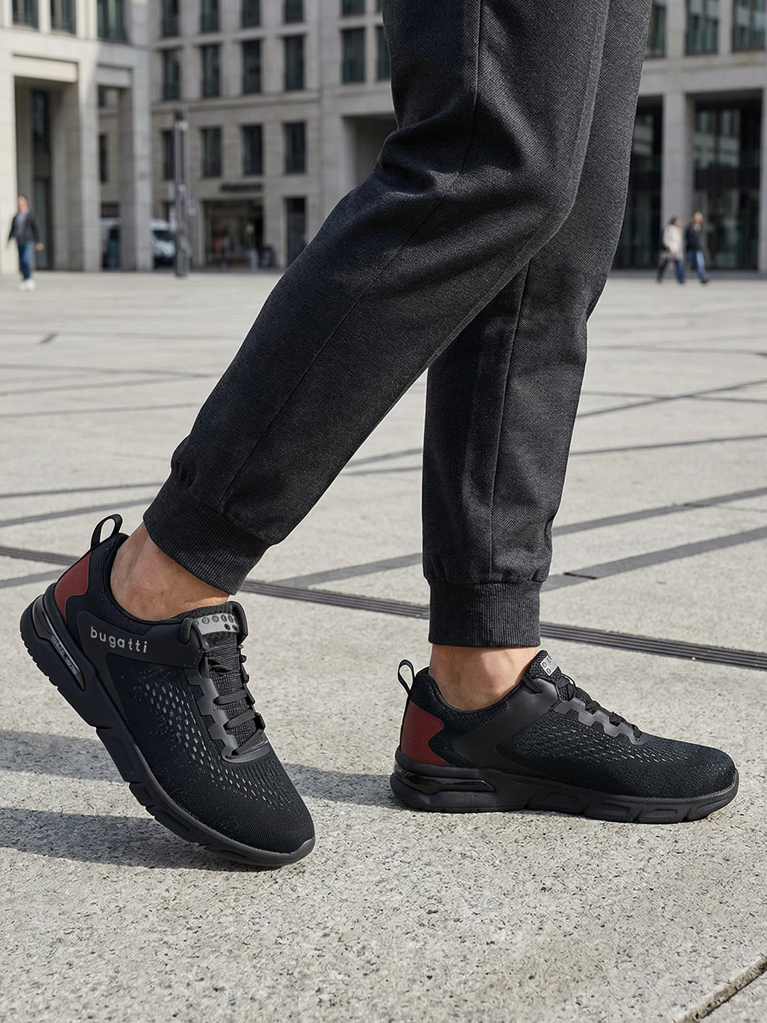 Lifestyle close-up of black bugatti Nubola comfort sneakers, with mesh texture and red heel accent, worn with dark gray sweatpants.
