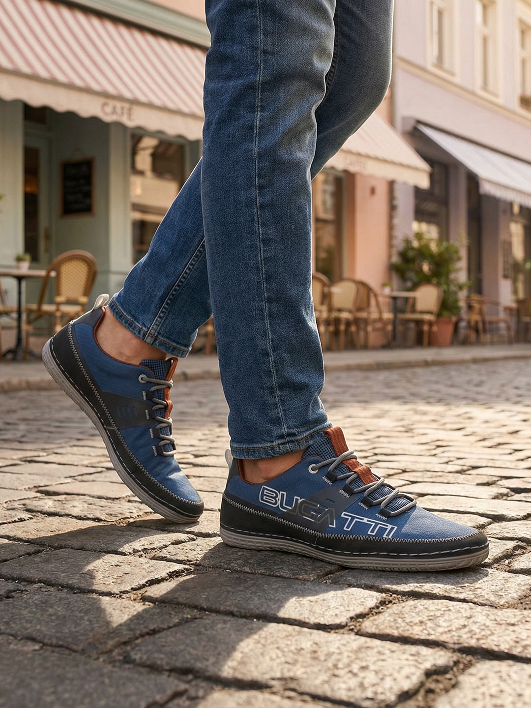 Lifestyle close-up of a person wearing blue denim jeans and bugatti Bimini blue low-top sneakers on a cobblestone street.