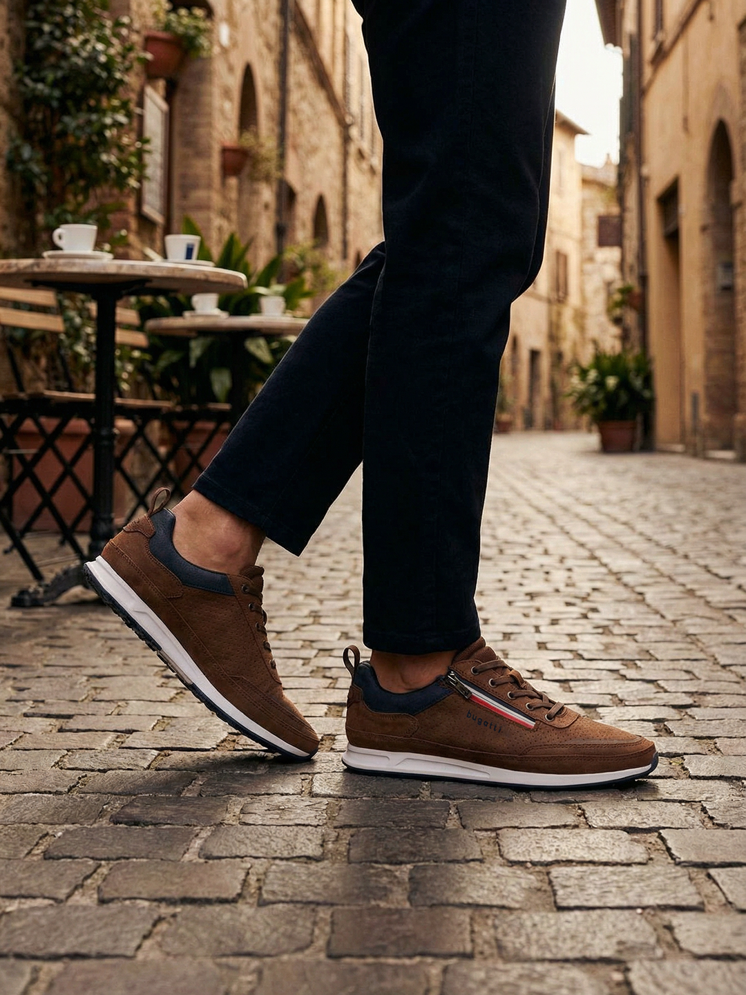 Lifestyle side view of the bugatti Ross Cognac Comfort Mid Top Sneakers, with blue accent and zipper detail, worn with black pants.
