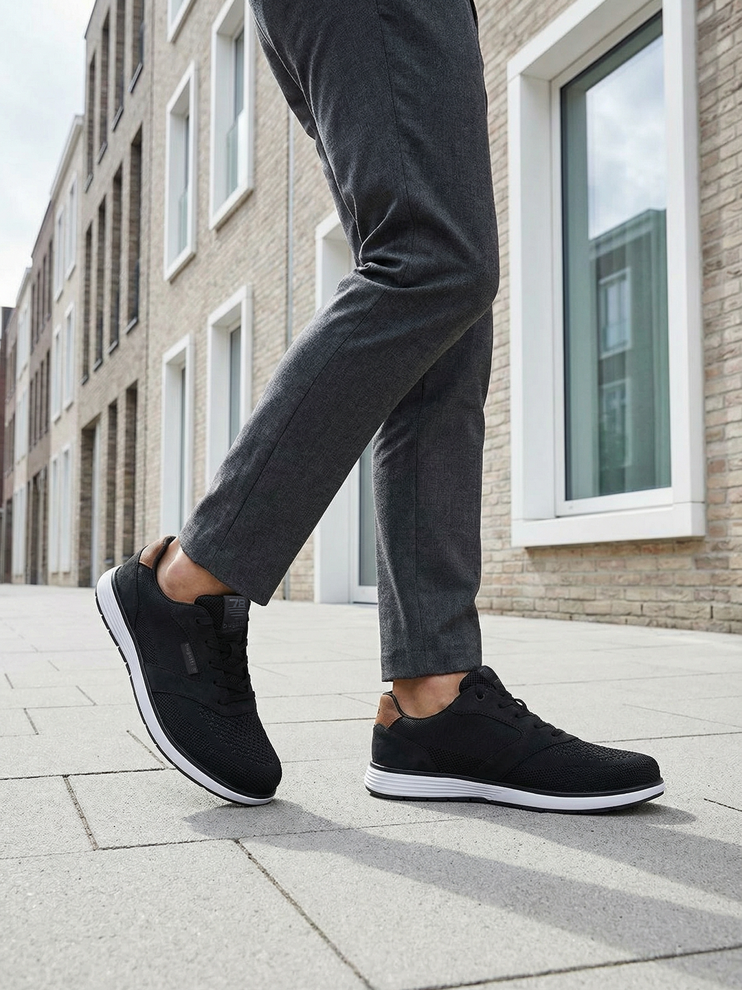 bugatti Baldini black sneakers, side view; low-cut with a white sole, worn with gray dress pants.