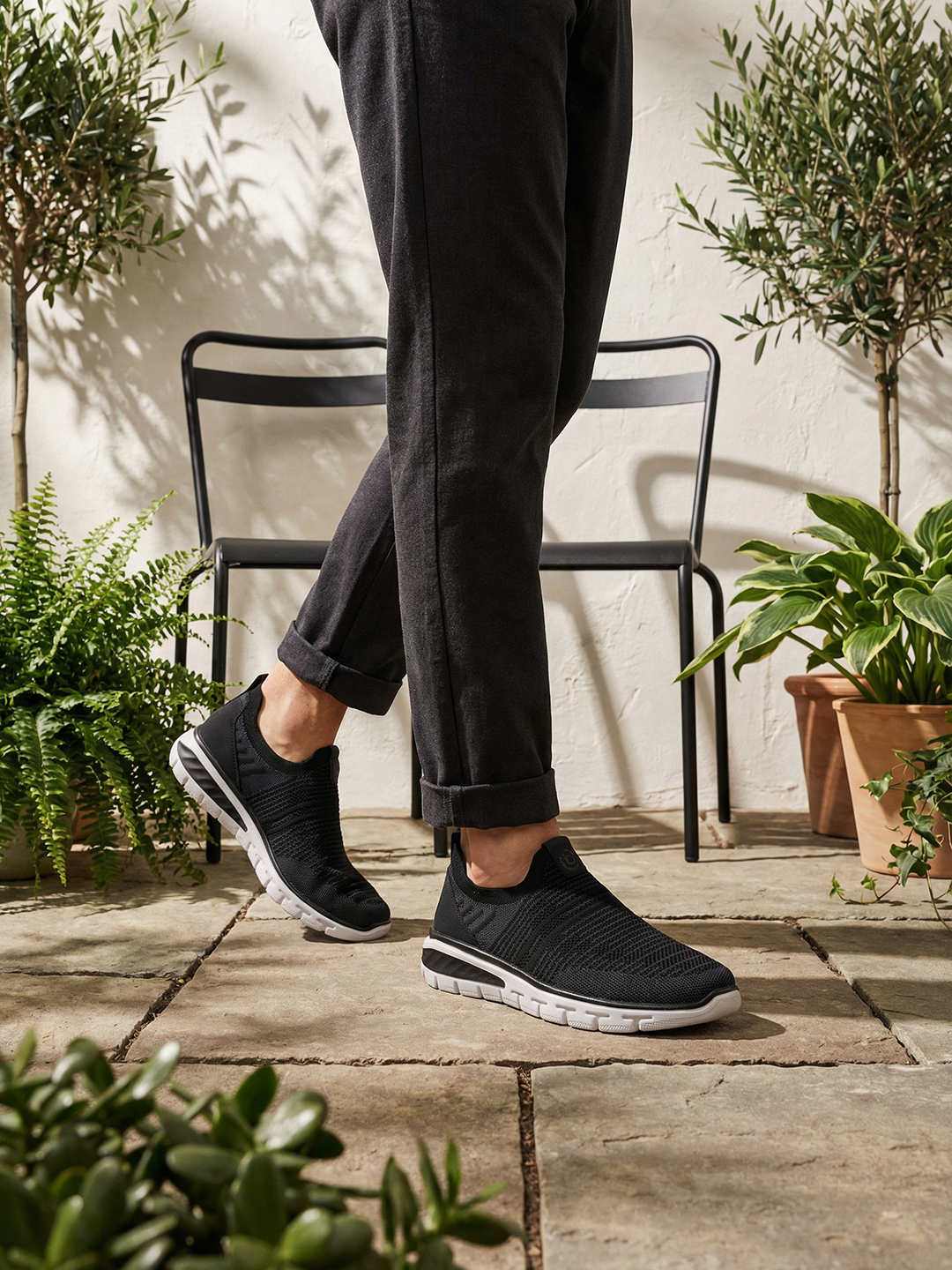Lifestyle image of bugatti Yucatan black comfort sneakers, with white soles, worn with cuffed black pants.
