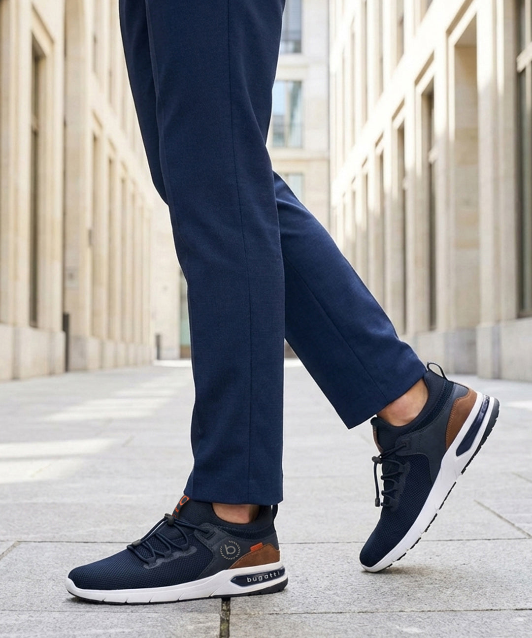 Lifestyle shot of bugatti Numbis dark blue athleisure sneakers with brown accents and white soles, paired with navy pants.
