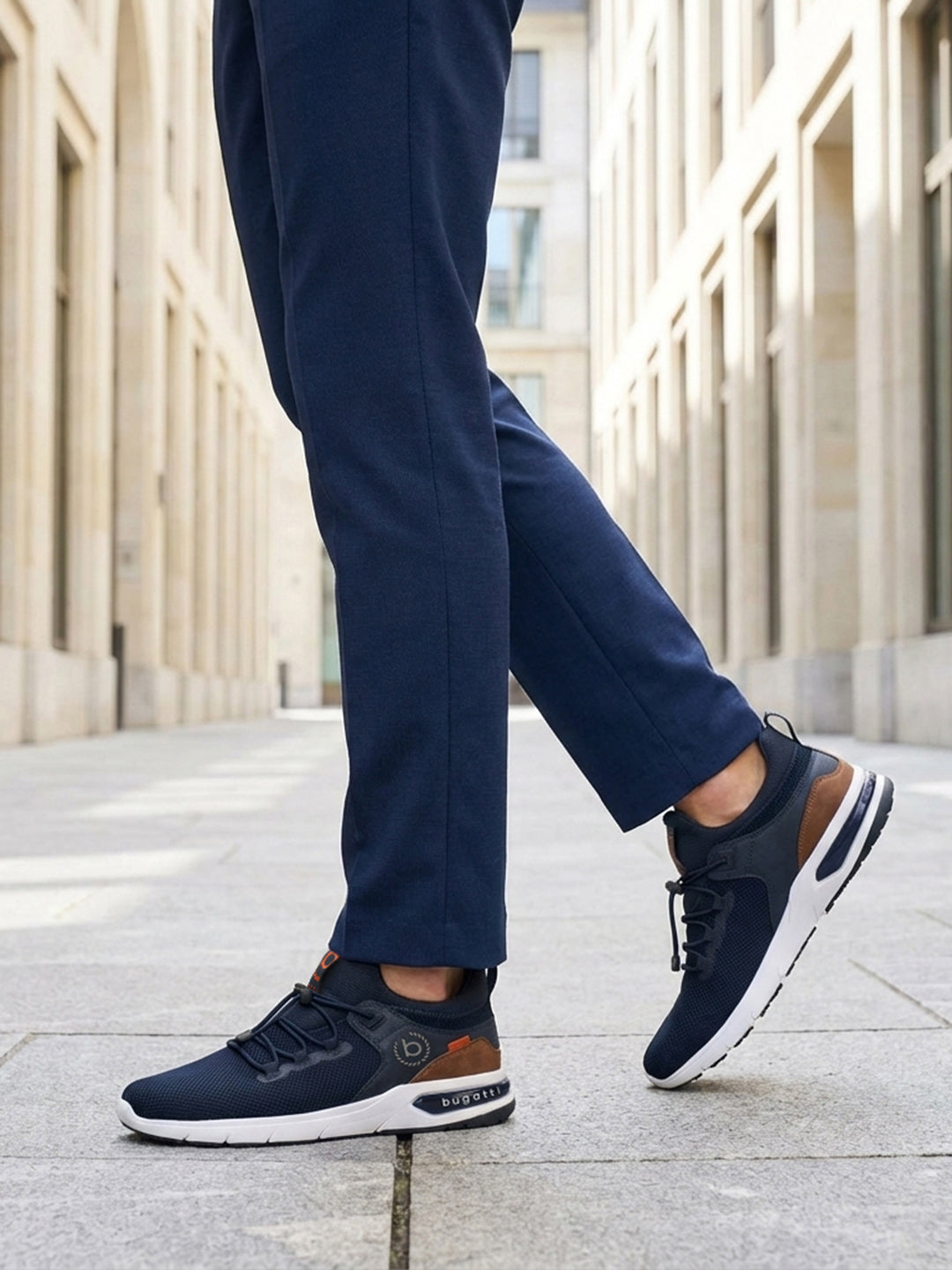 Lifestyle shot of bugatti Numbis dark blue athleisure sneakers with brown accents and white soles, paired with navy pants.

