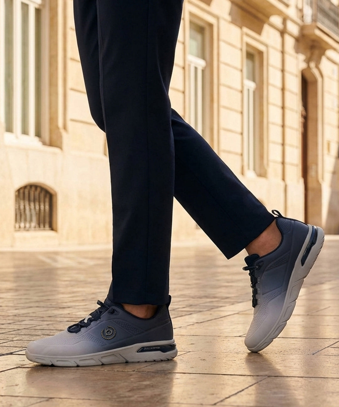 Lifestyle shot of bugatti Nubola light grey athleisure sneakers, with blue detailing, being worn with dark slacks.
