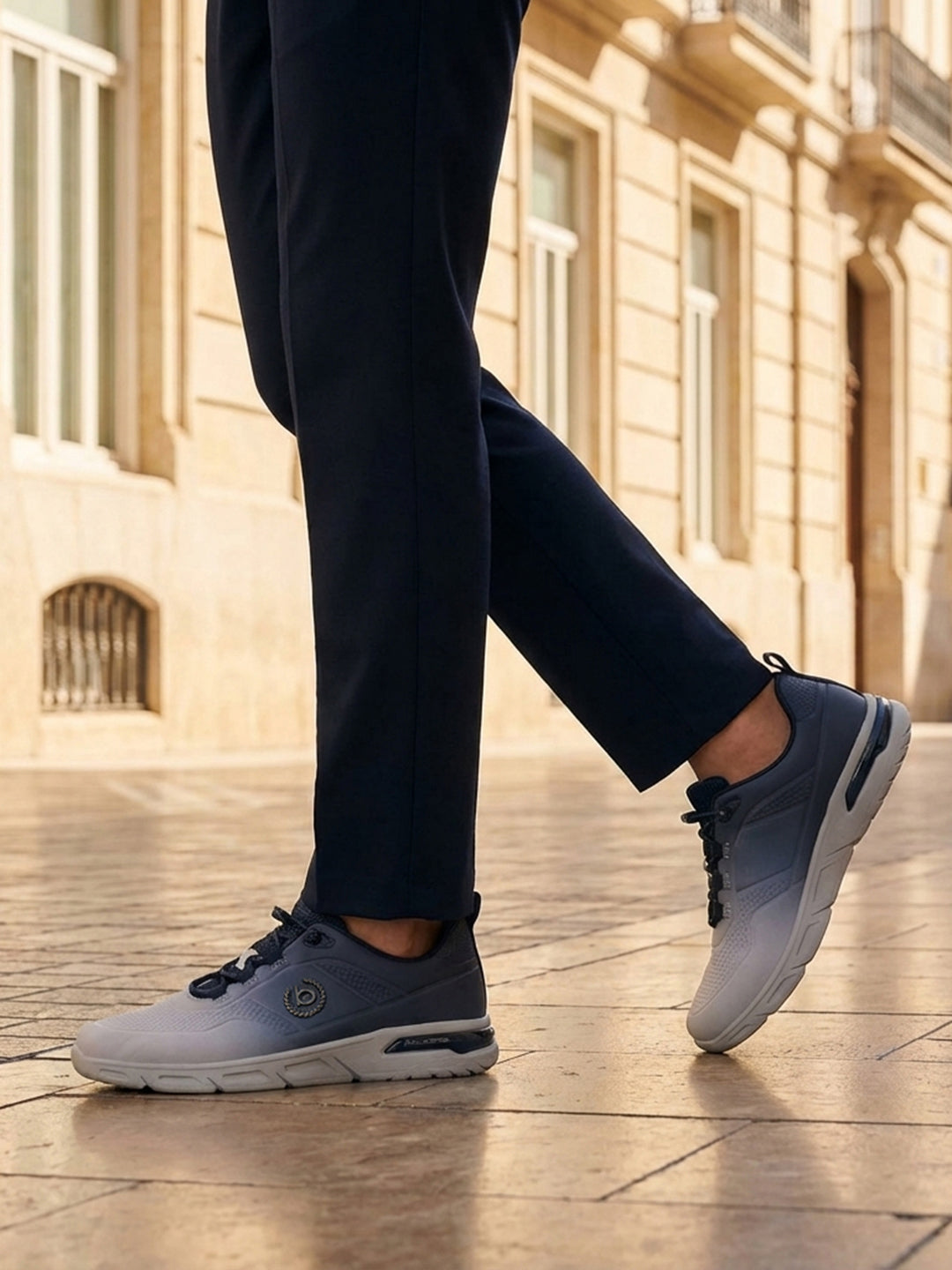 Lifestyle shot of bugatti Nubola light grey athleisure sneakers, with blue detailing, being worn with dark slacks.
