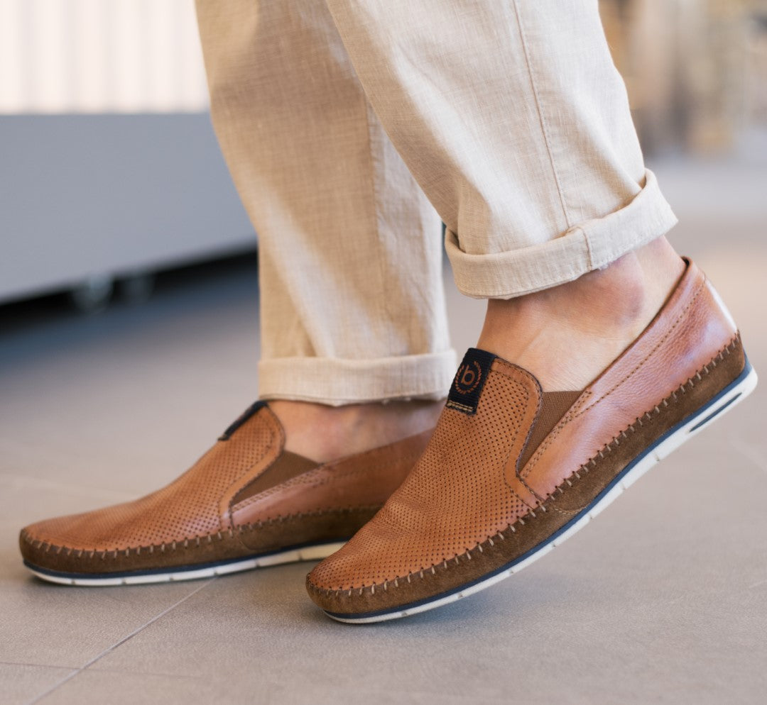 Cognac leather slip-on shoes by bugatti, with perforated texture and dark brown accents, worn with beige pants.